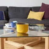 Table basse en verre et bois, avec un panier jaune, des tasses et des magazines. La hauteur de la table basse est idéale pour le canapé.