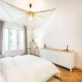 Un lustre design illumine cette chambre moderne. L'éclairage chambre est complété par une lampe sur trépied et une lumière douce émanant de la fenêtre.