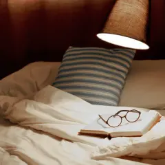 Un livre ouvert avec des lunettes repose sur un lit défait, sous une lampe. La hauteur appliquée au lit suggère un moment de détente avant le sommeil.