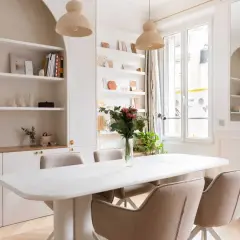 Salle à manger lumineuse avec table blanche, chaises beiges et un luminaire gradable suspendu. Étagères remplies de livres et d'objets décoratifs.
