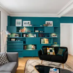 Un salon sombre avec une grande bibliothèque turquoise remplie de livres et d'objets décoratifs. Un canapé gris et un fauteuil noir complètent l'ensemble.