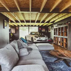 Un garage transformé en chambre avec une voiture de sport blanche, un grand canapé gris et une table basse rustique.