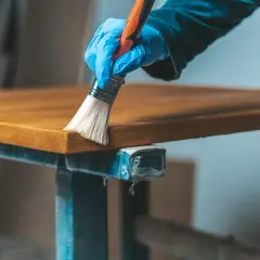 Main tenant, une main gantée applique un produit sur un plateau en bois vernis. Une étape pour comment éclaircir un bois foncé vernis.