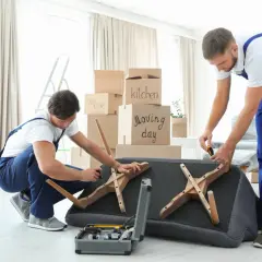 Deux déménageurs démontent un canapé fixe, entourés de cartons de déménagement. Ils utilisent des outils pour retirer les pieds en bois.