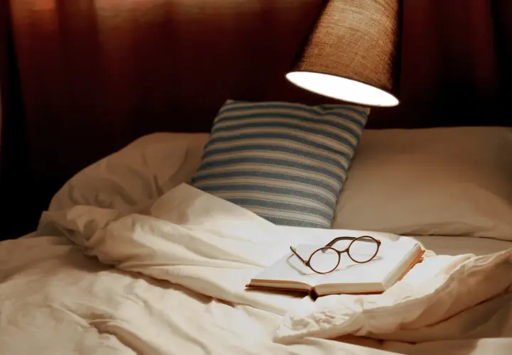 Un livre ouvert avec des lunettes repose sur un lit défait, sous une lampe. La hauteur appliquée au lit suggère un moment de détente avant le sommeil.