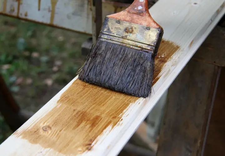 Application d'une couche de lasure sur une planche de bois avec un pinceau. Le bois prend une teinte dorée.