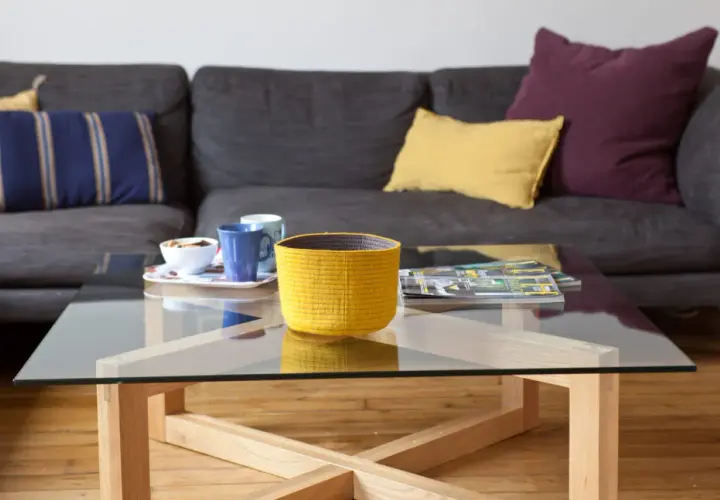 Table basse en verre et bois, avec un panier jaune, des tasses et des magazines. La hauteur de la table basse est idéale pour le canapé.