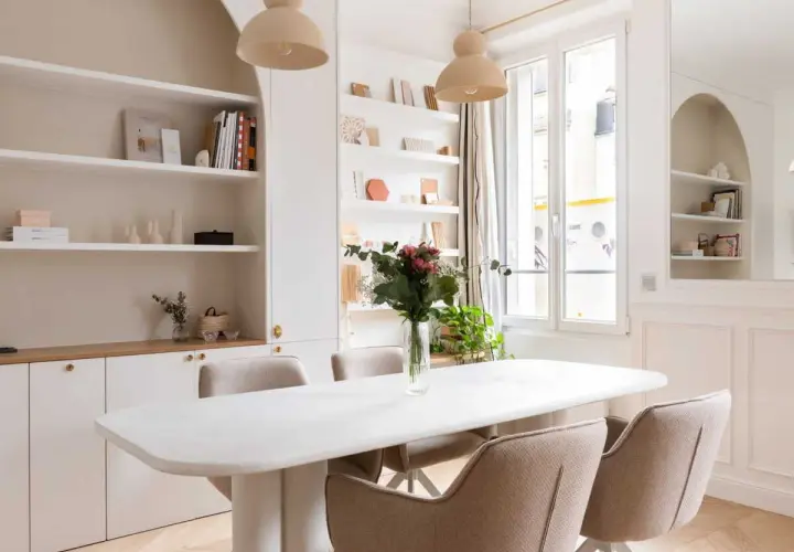 Salle à manger lumineuse avec table blanche, chaises beiges et un luminaire gradable suspendu. Étagères remplies de livres et d'objets décoratifs.