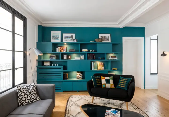 Un salon sombre avec une grande bibliothèque turquoise remplie de livres et d'objets décoratifs. Un canapé gris et un fauteuil noir complètent l'ensemble.