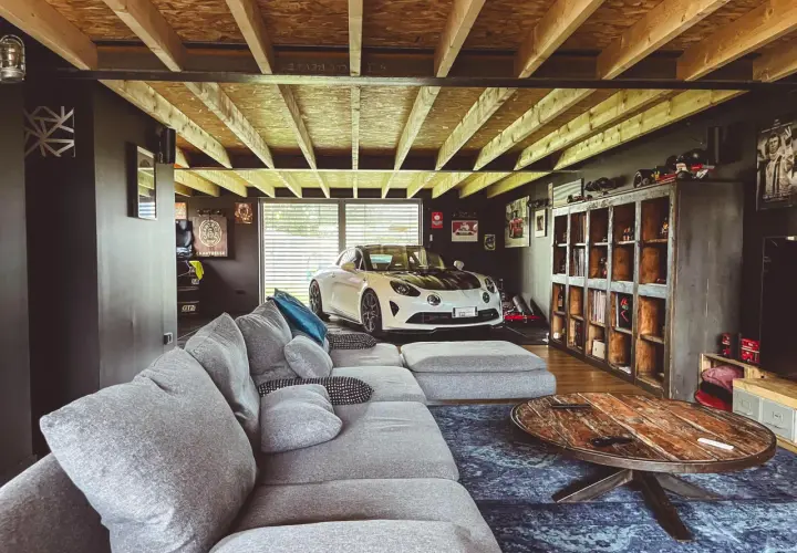 Un garage transformé en chambre avec une voiture de sport blanche, un grand canapé gris et une table basse rustique.