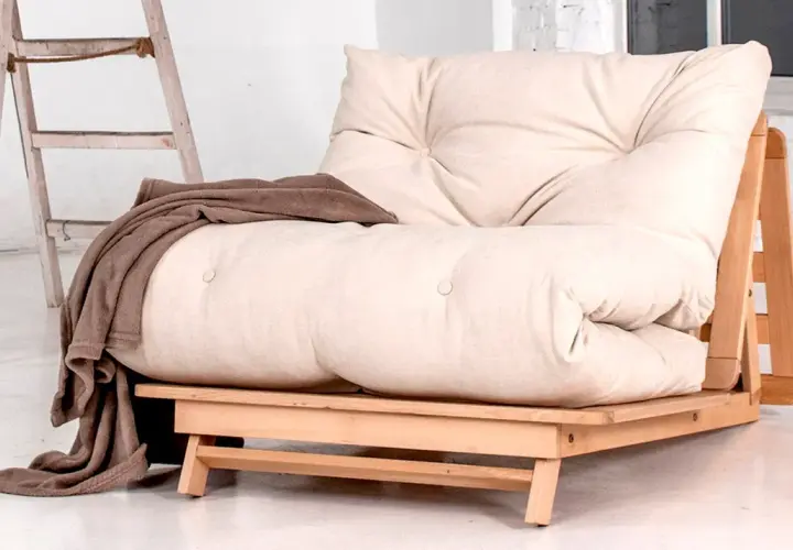 Un canapé futon beige avec une couverture marron drapée. Idéal pour savoir comment rendre un canapé plus confortable.