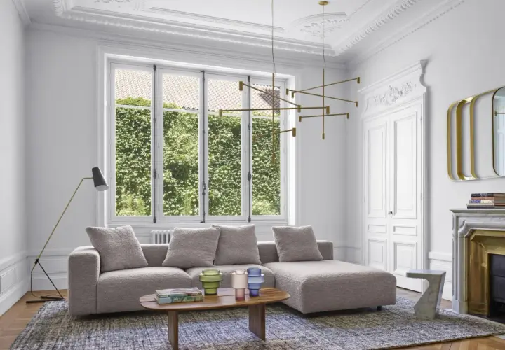 Salon moderne avec un grand canapé gris moelleux, une table basse en bois, un lustre doré et une cheminée ornée.
