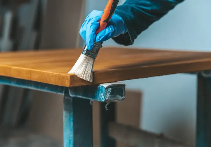 Main tenant, une main gantée applique un produit sur un plateau en bois vernis. Une étape pour comment éclaircir un bois foncé vernis.