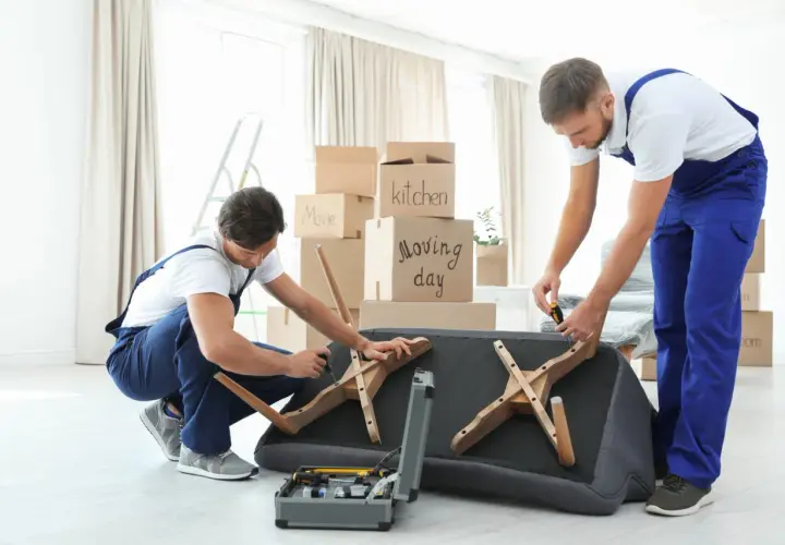 Deux déménageurs démontent un canapé fixe, entourés de cartons de déménagement. Ils utilisent des outils pour retirer les pieds en bois.