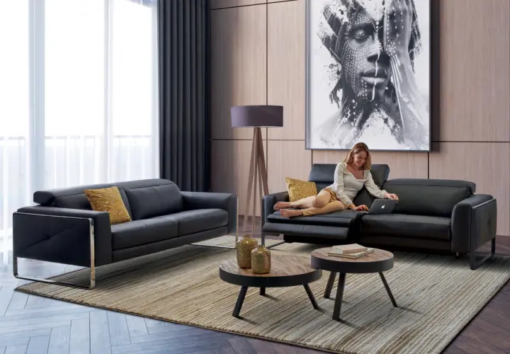 Salon moderne avec canapé noir, femme sur un fauteuil, tableau abstrait et tables basses.