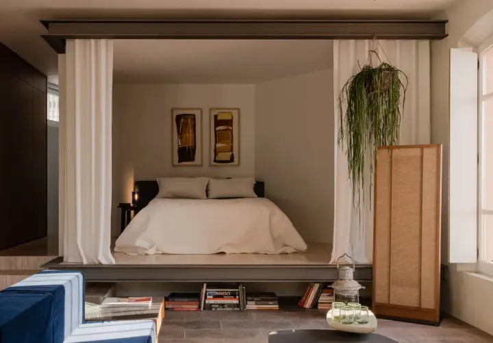 Chambre avec estrade, séparée par des rideaux blancs. Un lit douillet, des tableaux abstraits et une plante suspendue créent une atmosphère paisible.