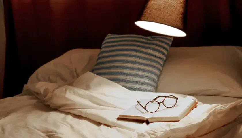 Un livre ouvert avec des lunettes repose sur un lit défait, sous une lampe. La hauteur appliquée au lit suggère un moment de détente avant le sommeil.