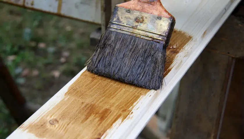 Application d'une couche de lasure sur une planche de bois avec un pinceau. Le bois prend une teinte dorée.