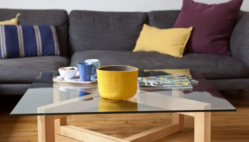 Table basse en verre et bois, avec un panier jaune, des tasses et des magazines. La hauteur de la table basse est idéale pour le canapé.