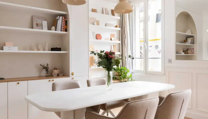 Salle à manger lumineuse avec table blanche, chaises beiges et un luminaire gradable suspendu. Étagères remplies de livres et d'objets décoratifs.
