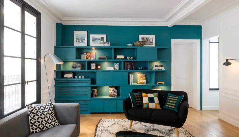 Un salon sombre avec une grande bibliothèque turquoise remplie de livres et d'objets décoratifs. Un canapé gris et un fauteuil noir complètent l'ensemble.