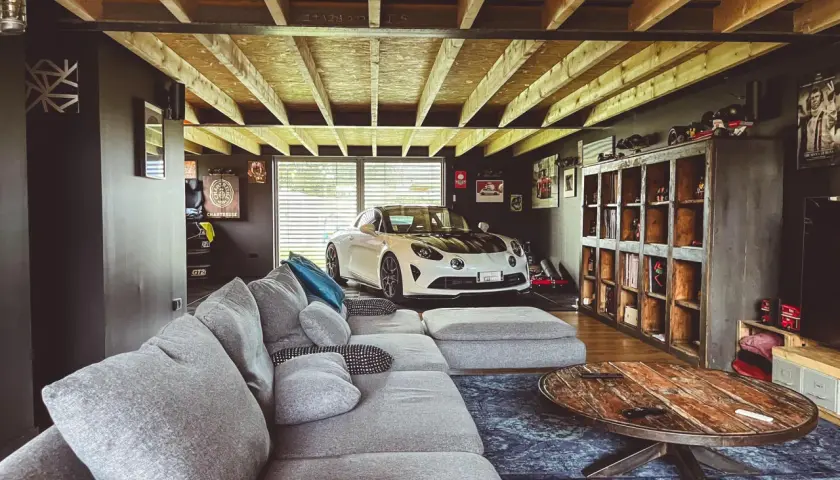 Un garage transformé en chambre avec une voiture de sport blanche, un grand canapé gris et une table basse rustique.