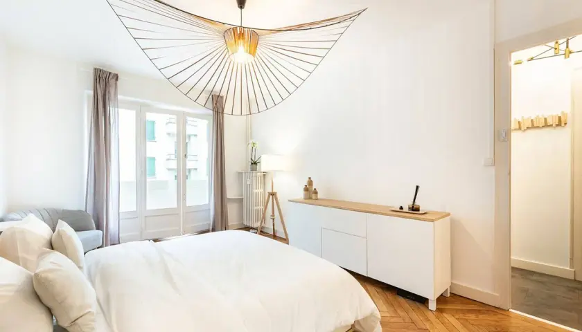 Un lustre design illumine cette chambre moderne. L'éclairage chambre est complété par une lampe sur trépied et une lumière douce émanant de la fenêtre.