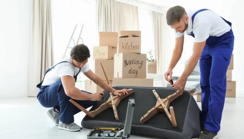 Deux déménageurs démontent un canapé fixe, entourés de cartons de déménagement. Ils utilisent des outils pour retirer les pieds en bois.