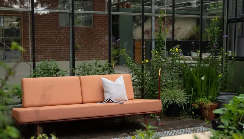 Un canapé décoloré par le soleil, entouré de verdure luxuriante dans une serre. Une coussin blanc repose sur le siège.