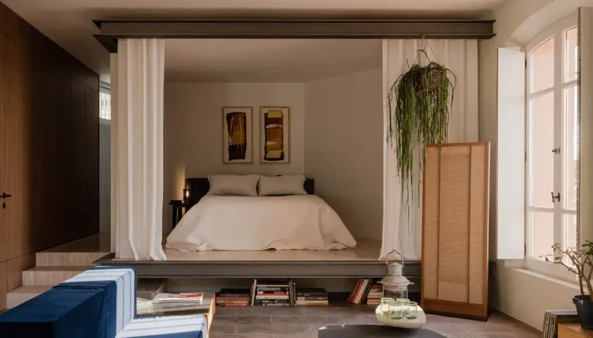 Chambre avec estrade, séparée par des rideaux blancs. Un lit douillet, des tableaux abstraits et une plante suspendue créent une atmosphère paisible.