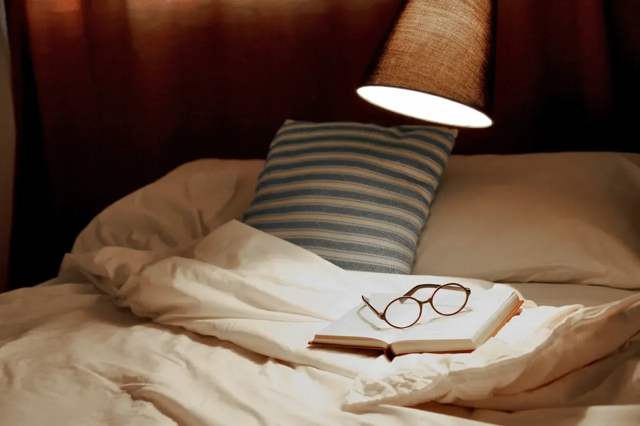 Un livre ouvert avec des lunettes repose sur un lit défait, sous une lampe. La hauteur appliquée au lit suggère un moment de détente avant le sommeil.