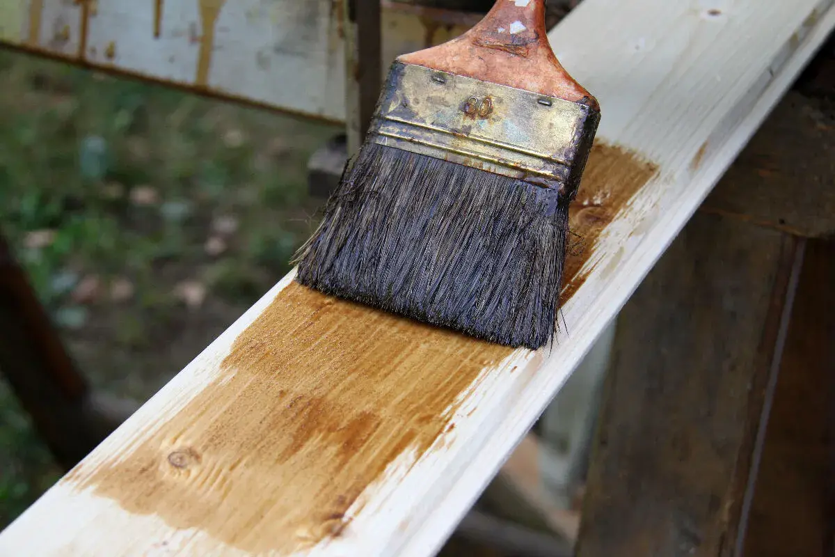 Application d'une couche de lasure sur une planche de bois avec un pinceau. Le bois prend une teinte dorée.