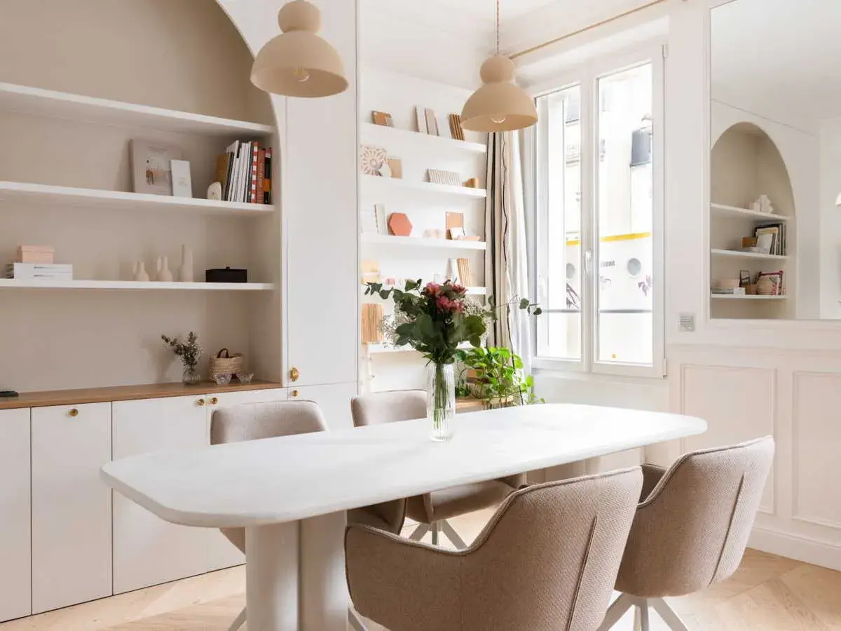 Salle à manger lumineuse avec table blanche, chaises beiges et un luminaire gradable suspendu. Étagères remplies de livres et d'objets décoratifs.