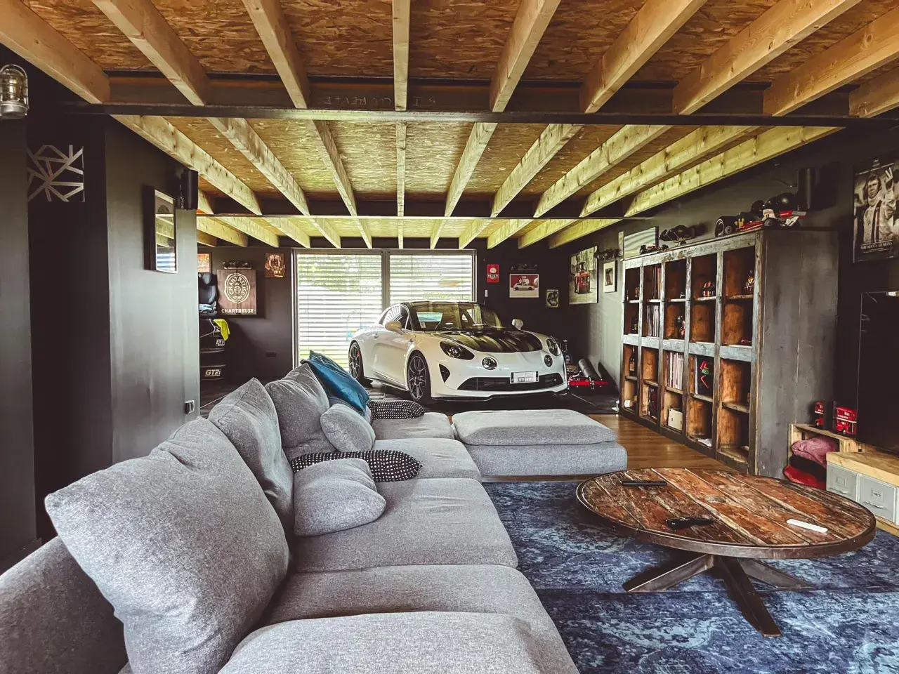 Un garage transformé en chambre avec une voiture de sport blanche, un grand canapé gris et une table basse rustique.