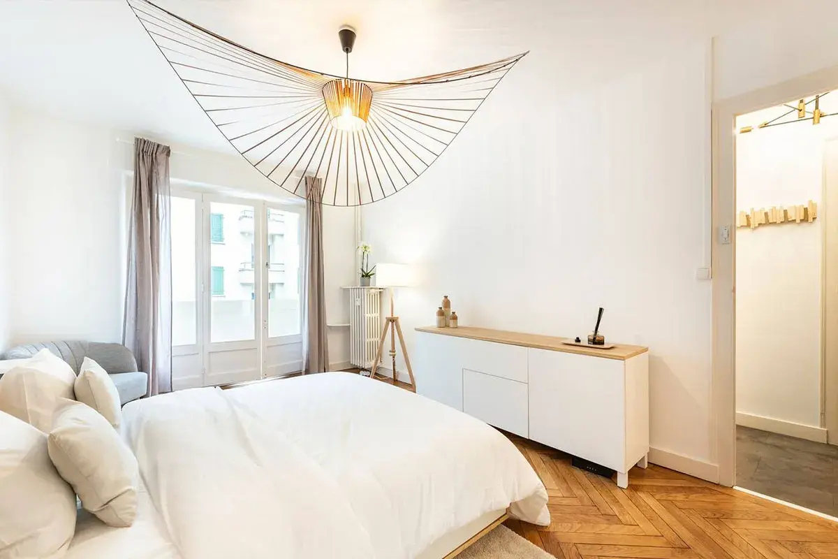 Un lustre design illumine cette chambre moderne. L'éclairage chambre est complété par une lampe sur trépied et une lumière douce émanant de la fenêtre.