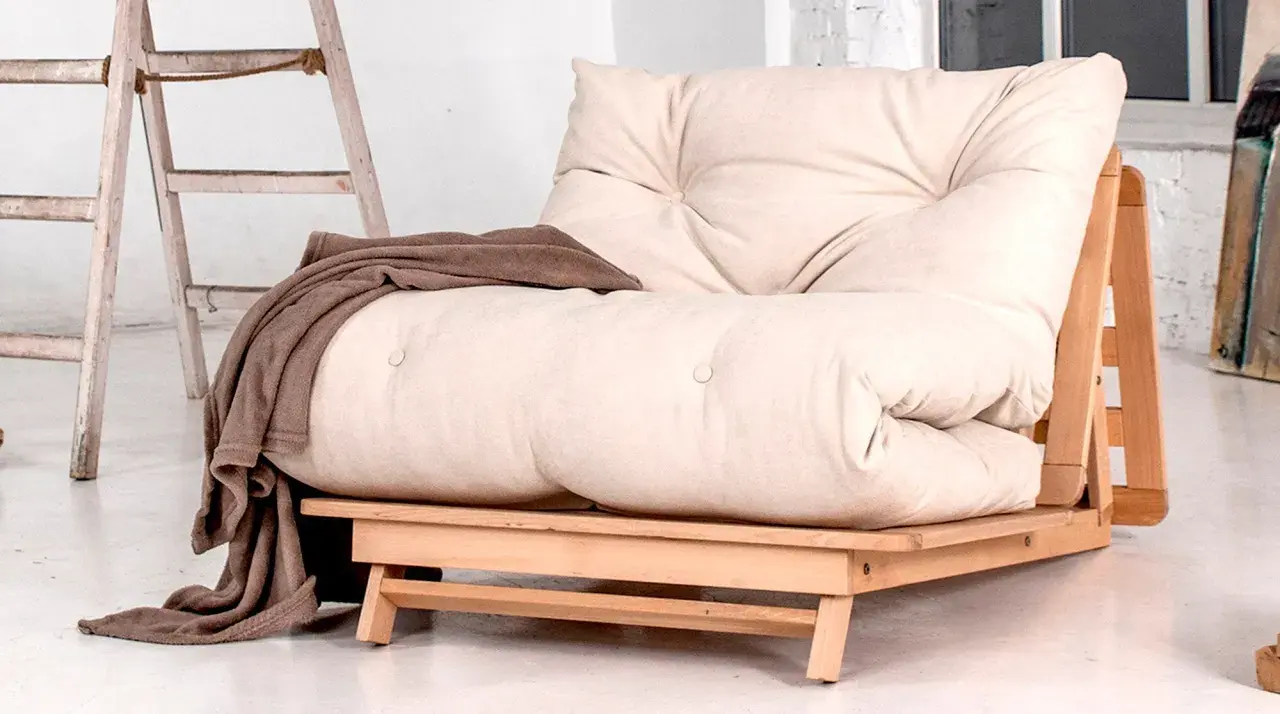 Un canapé futon beige avec une couverture marron drapée. Idéal pour savoir comment rendre un canapé plus confortable.