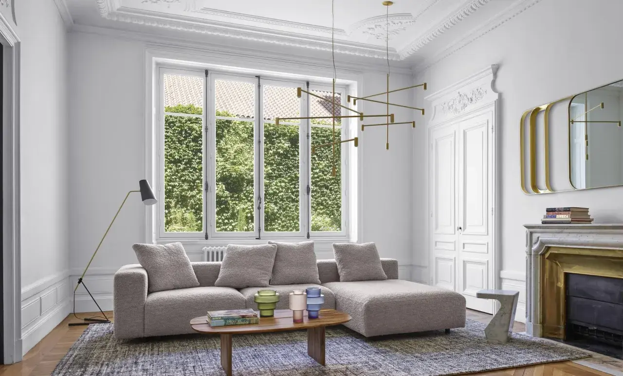 Salon moderne avec un grand canapé gris moelleux, une table basse en bois, un lustre doré et une cheminée ornée.