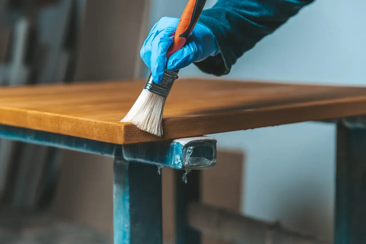Main tenant, une main gantée applique un produit sur un plateau en bois vernis. Une étape pour comment éclaircir un bois foncé vernis.