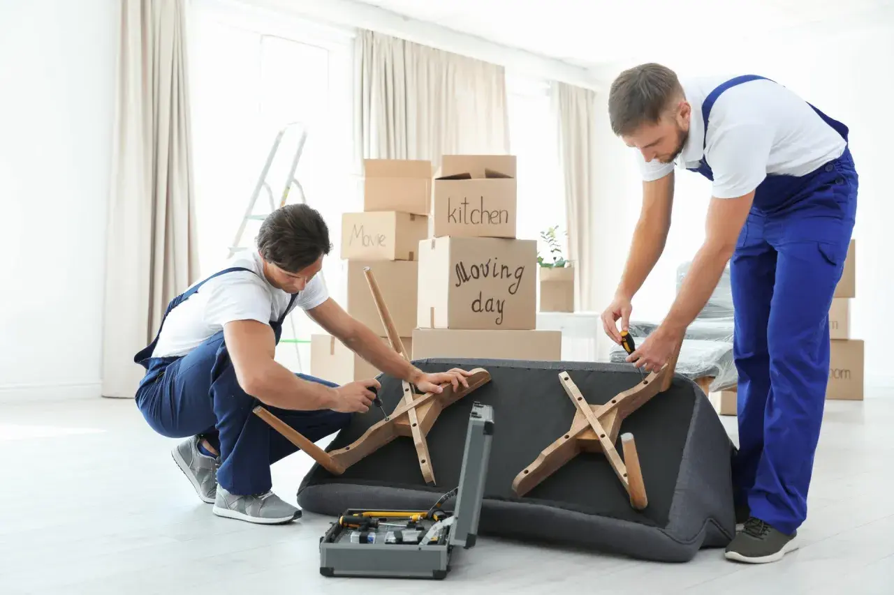 Deux déménageurs démontent un canapé fixe, entourés de cartons de déménagement. Ils utilisent des outils pour retirer les pieds en bois.