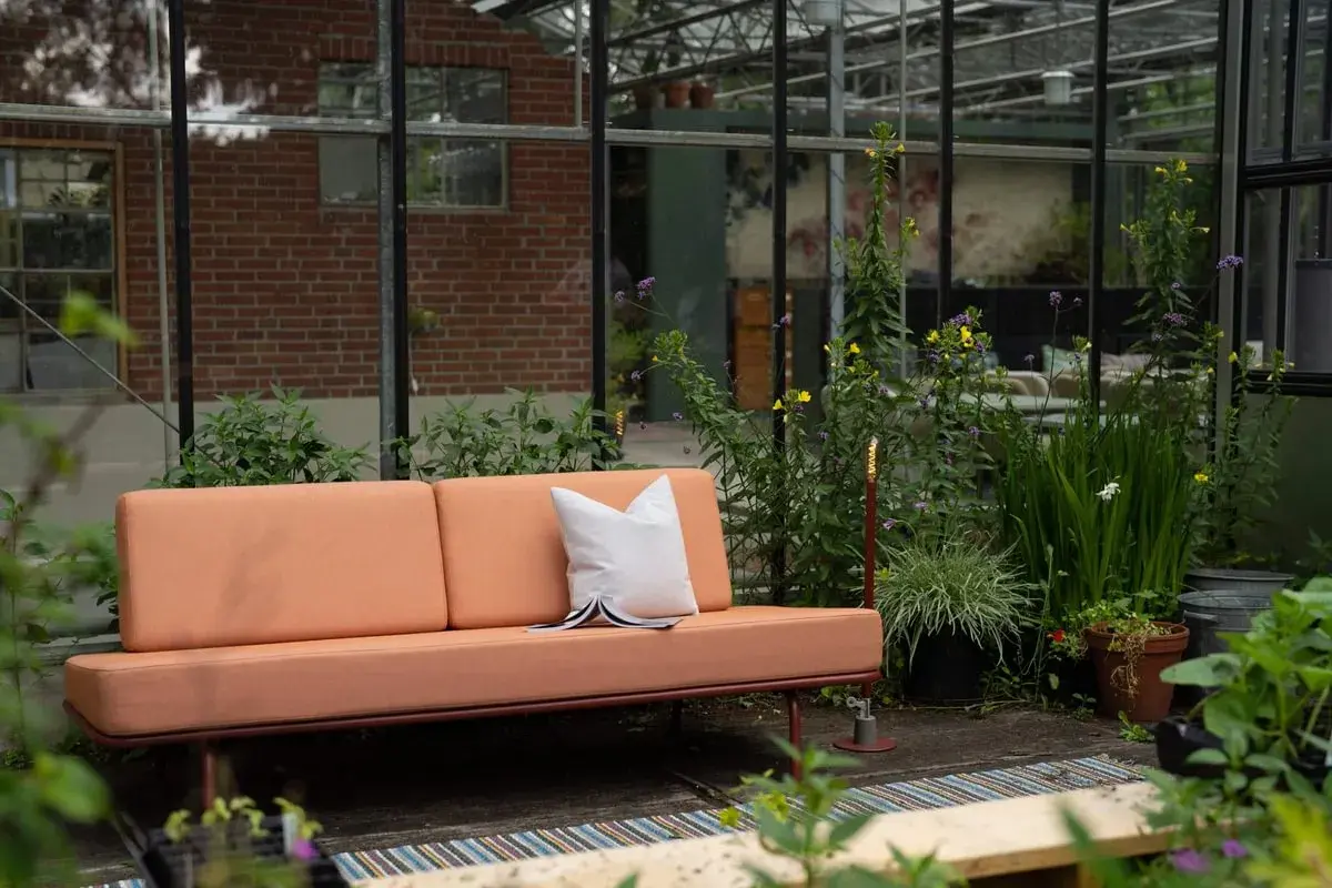 Un canapé décoloré par le soleil, entouré de verdure luxuriante dans une serre. Une coussin blanc repose sur le siège.