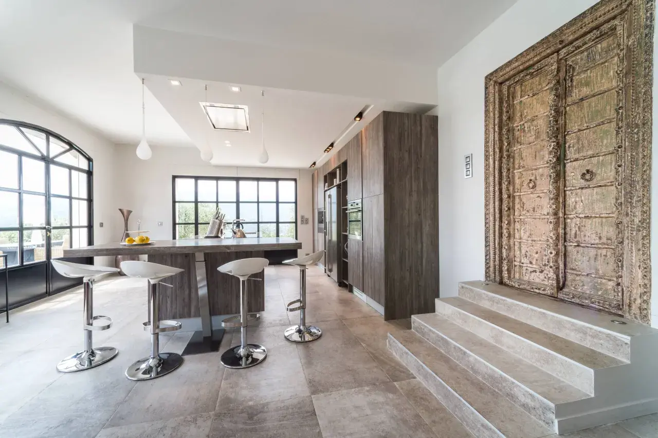 Cuisine moderne avec îlot central, tabourets chromés et une porte ancienne ornée. Un mélange audacieux de style Louis Philippe et contemporain.