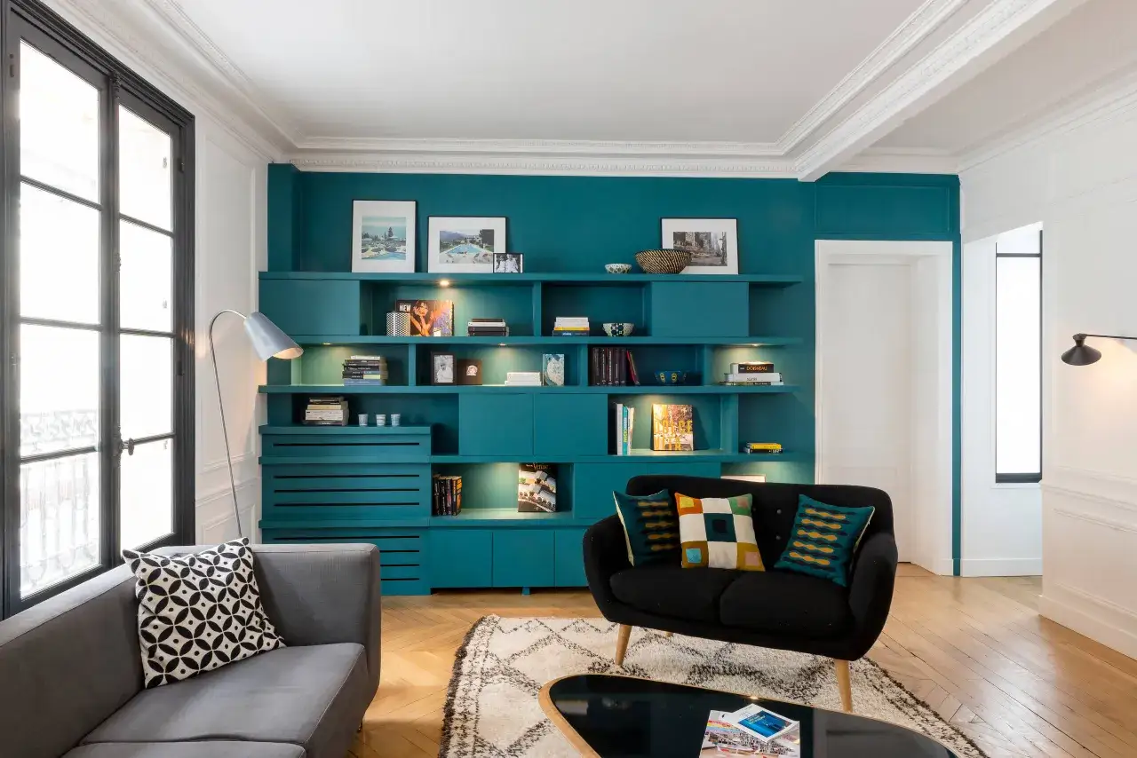 Un salon sombre avec une grande bibliothèque turquoise remplie de livres et d'objets décoratifs. Un canapé gris et un fauteuil noir complètent l'ensemble.