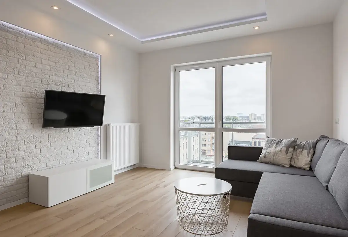Salon moderne avec TV murale &agrave; bonne hauteur, canap&eacute; gris, table basse et grande fen&ecirc;tre offrant une vue sur la ville.