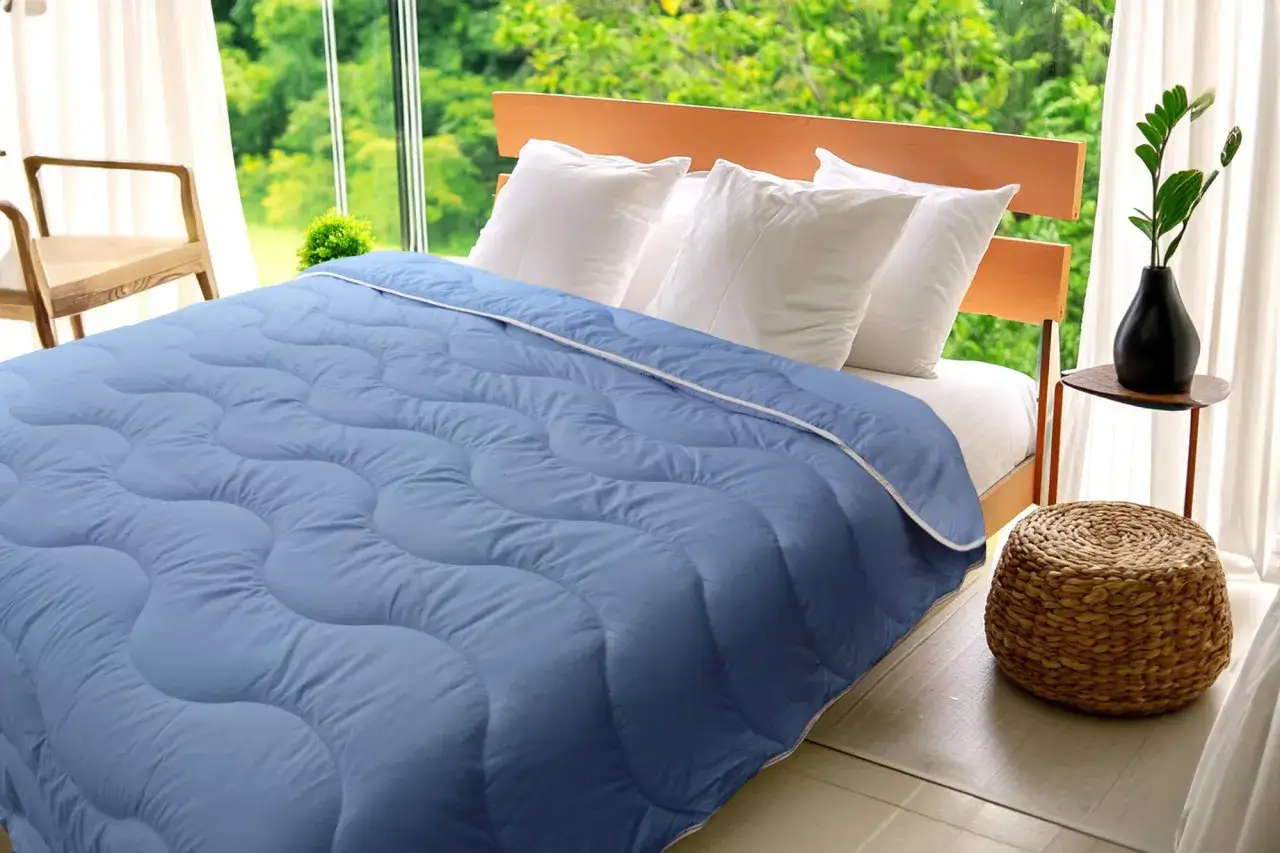 Une couette bleue légère, parfaite pour l'été, est étalée sur un lit. La fenêtre laisse voir une végétation luxuriante.