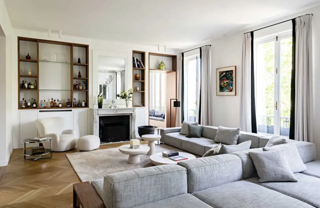 Salon moderne et lumineux, parfait pour le home staging. Canapé gris, cheminée, étagères remplies de bouteilles et fauteuil blanc invitent à la détente.