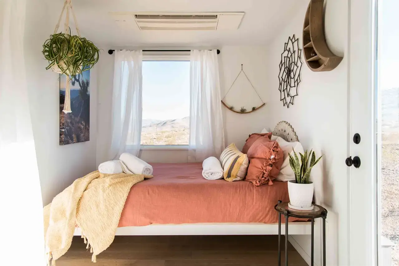 Une chambre d'amis cosy avec un lit orange, des coussins texturés, une couverture jaune et une plante verte. La fenêtre offre une vue sur un paysage désertique.