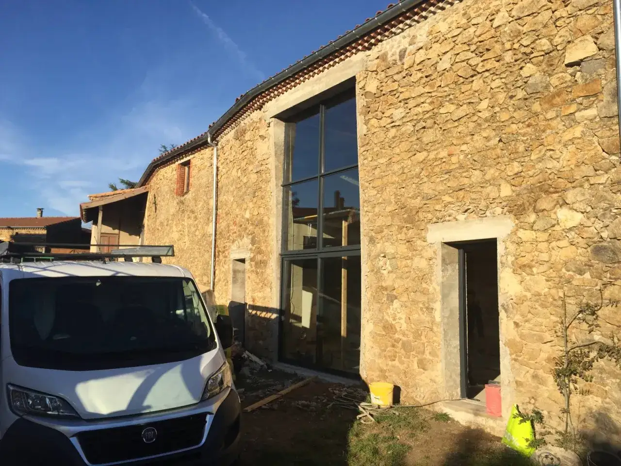 Une grande baie vitrée et une porte créent une nouvelle ouverture façade maison en pierre. Un fourgon de chantier est garé devant.