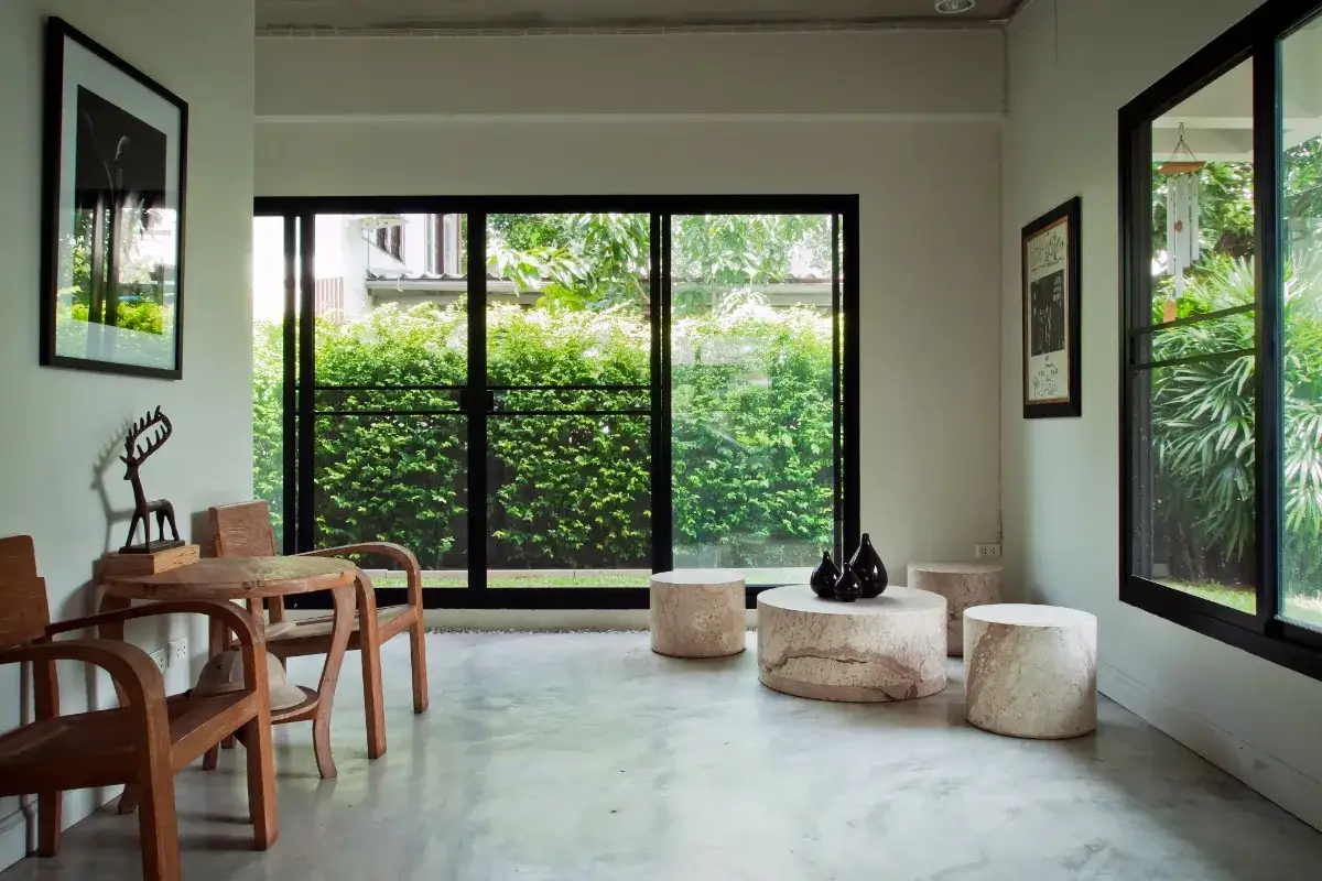 Salon moderne avec un sol en béton ciré, des meubles en bois, de grandes fenêtres donnant sur un jardin luxuriant et des œuvres d'art.