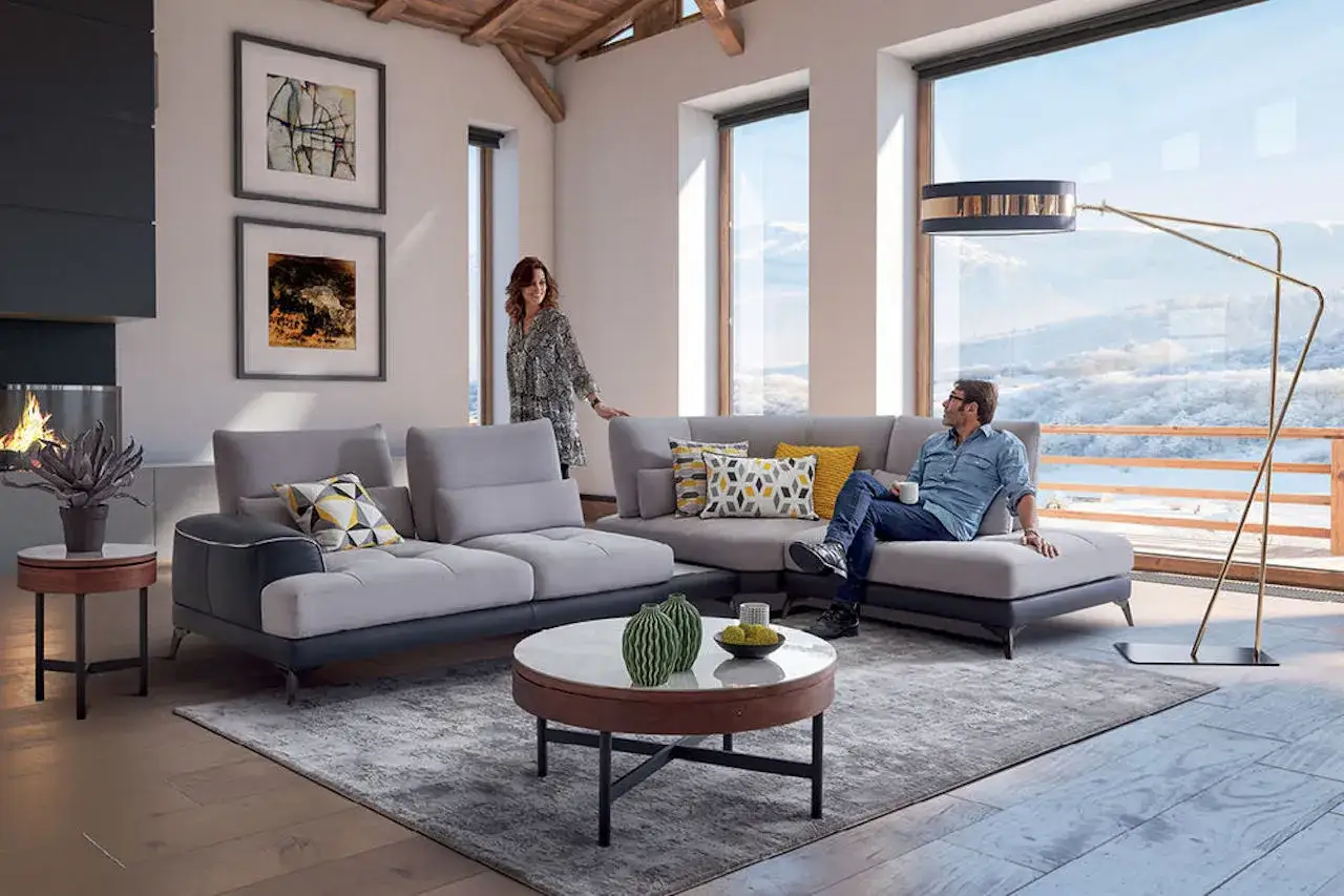 Un couple profite d'un salon moderne avec un grand canap&eacute; d'angle, une chemin&eacute;e et une vue panoramique sur la montagne.