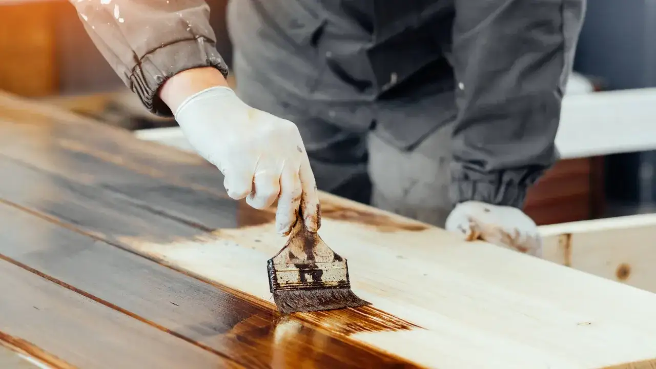 Main d'un artisan gant&eacute; appliquant une teinture fonc&eacute;e pour repeindre une table en bois, transformant le meuble.